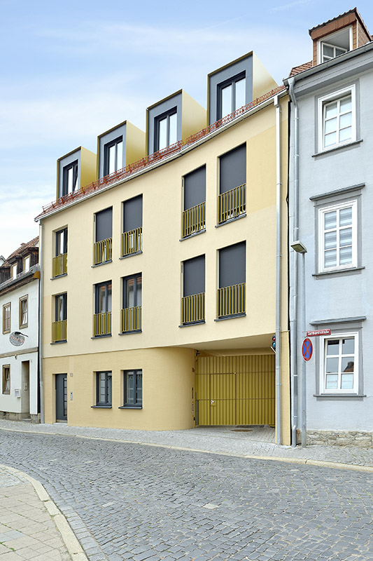 In Gelbtönen gestrichene Gebäudefassade in einer Häuserzeile, davor eine gepflasterte Straße