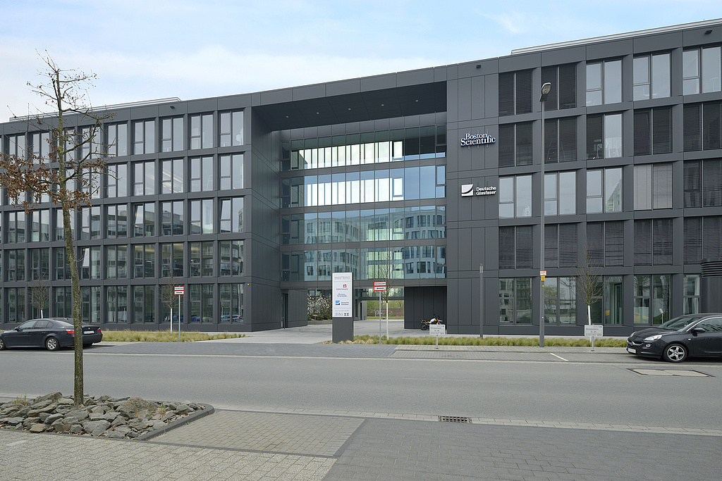 Verglaste Gebäudefront, in der Mitte verglaster Zugangsbereich, davor eine Straße mit geparkten Autos