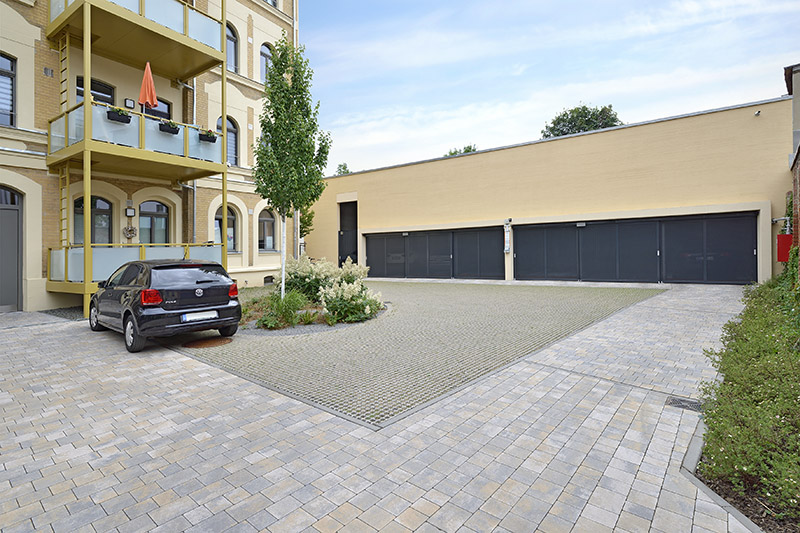 Links historisches Gebäude mit Anbaubalkonen, rechts flaches Gebäude mit mehreren Schiebetoren, davor ein großer gepflasterter Platz.